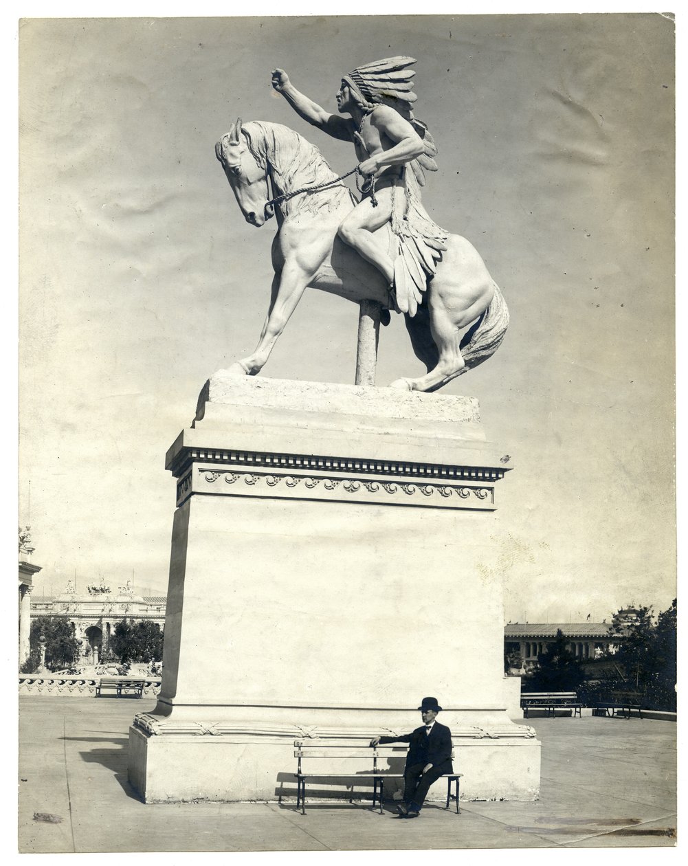 Vertical, sepia photograph showing a statue of an American Indian seated on a horse on top of a large pedestal. There is a man in a dark suit and hat seated on a bench at the pedestal