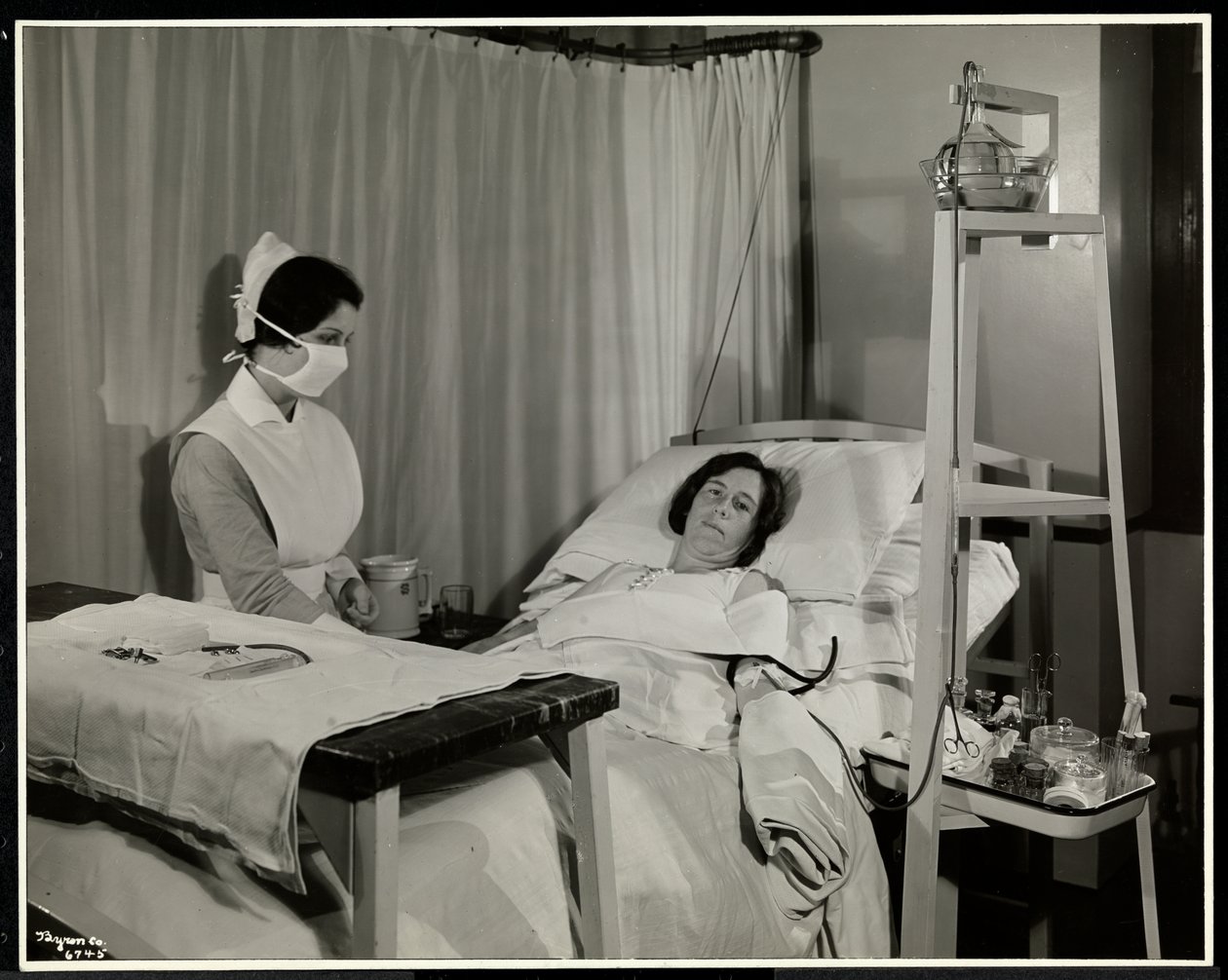En maskerad sjuksköterska med en patient i ett rum på Sloane Medical Hospital, Broadway och 168th Street, New York, 1932 (silvergelatintryck) av Byron Company