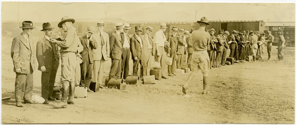 Horizontal, sepia photograph showing a group of men in civilian clothing, many of whom have luggage, standing in a line. Uniformed soldiers are walking along the line speaking with the men individually. Raildroad tracks and train cars can be seen in the ba av Carl Michel