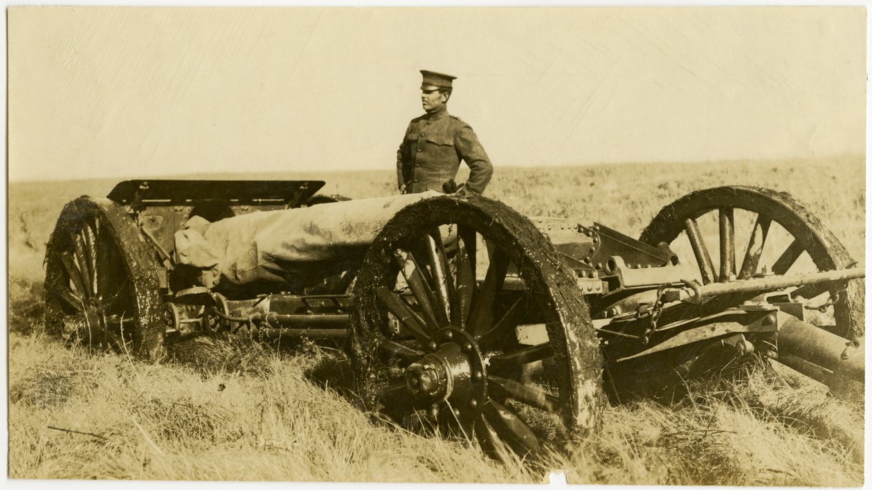Horizontal, sepia photograph showing a uniformed man standing next to an artillery gun on wheels in an open field av Carl Michel