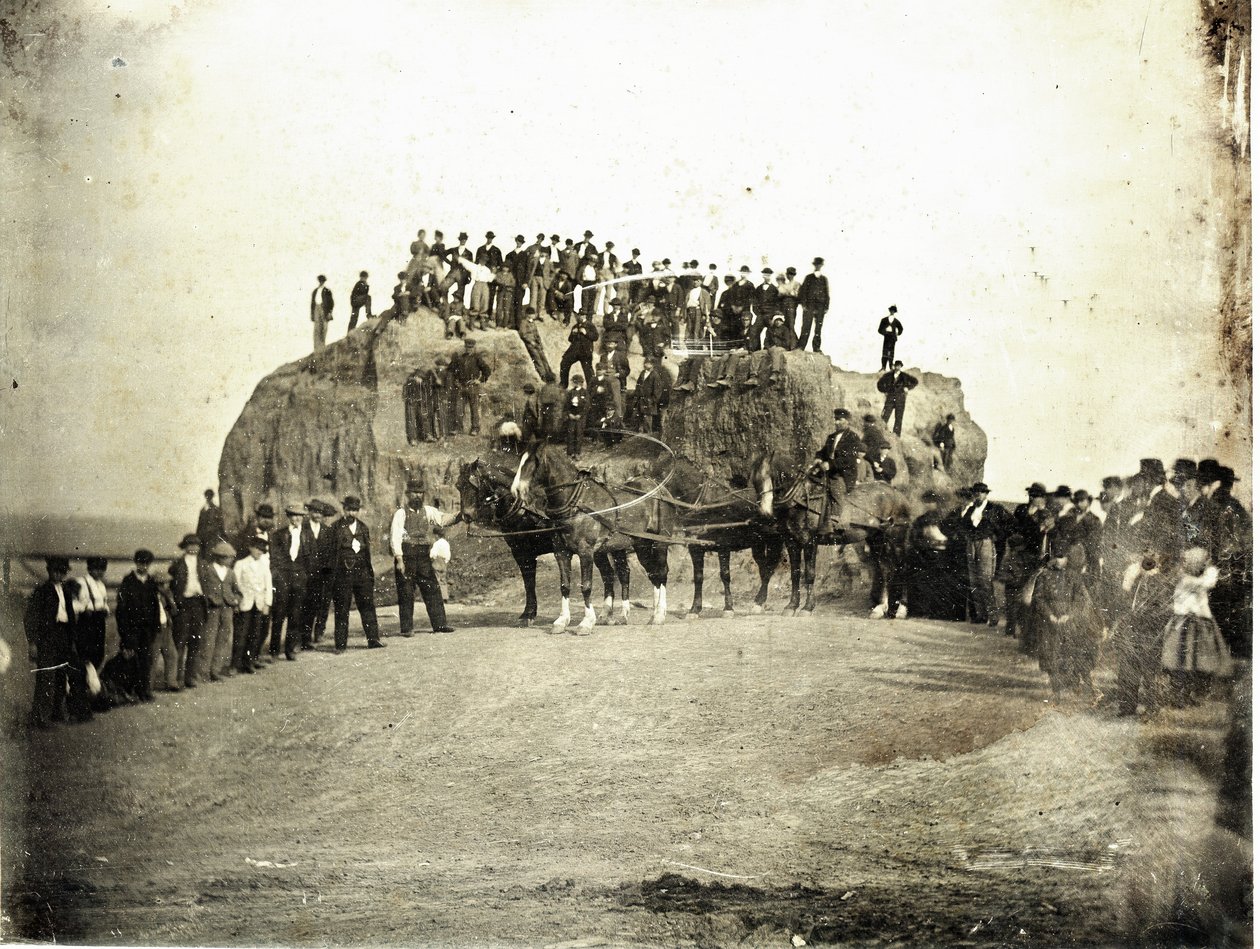 A crowd of men, children, and a few horses, stand on top of and in front of the Big Mound which is shown partially graded av Thomas M. Easterly