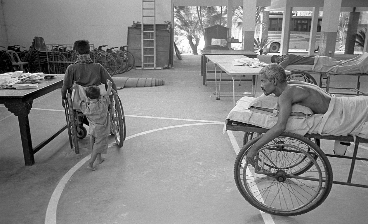 En fysiskt utmanad man i rullstol bärs av sin son; en annan man med samma svåra situation tittar på det på Center for the Rehabilitation of the Paralysed, CRP i Savar, Dhaka, Bangladesh. 2005. av Unbekannt Unbekannt