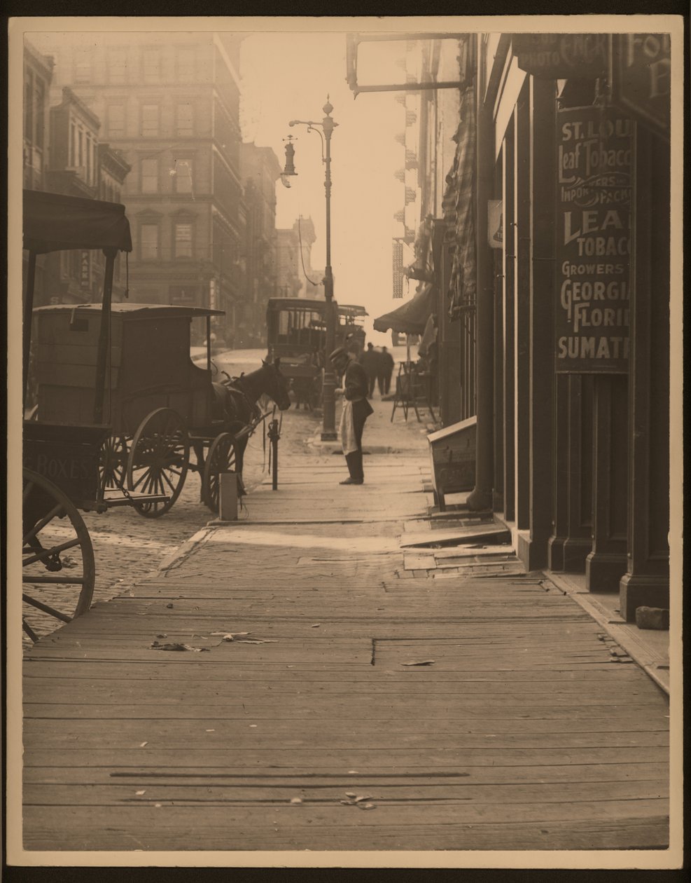 Vertical, black and white photograph of a horse-drawn vehicles parked against a wood plank sidewalk. Store fronts line the sidewalk and a man stands on the corner av William G. Swekosky